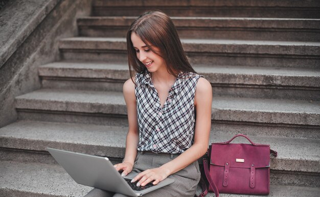 A cheerful student sits on the steps near the campus and dials on the laptop's keyboard