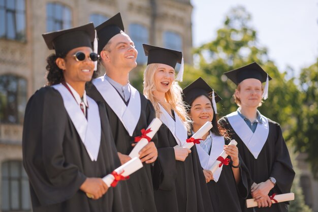 Cheerful students wearing graduation gowns standing next to each other