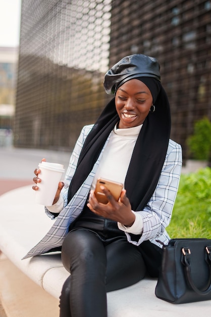 Cheerful stylish black woman in headscarf using smartphone in city