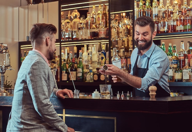 Веселый стильный брутальный бармен в рубашке и фартуке обслуживает клиентов на фоне барной стойки.