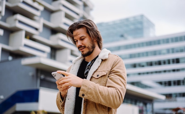 Cheerful stylish guy typing message and looking at the smartphone screen while walking at the city