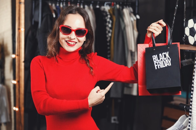 Cheerful successful happy young girl wearing sunglasses and red dresses showing black friday bag in fashion mall during shopping process concept of consumerism Black Friday sale rich life