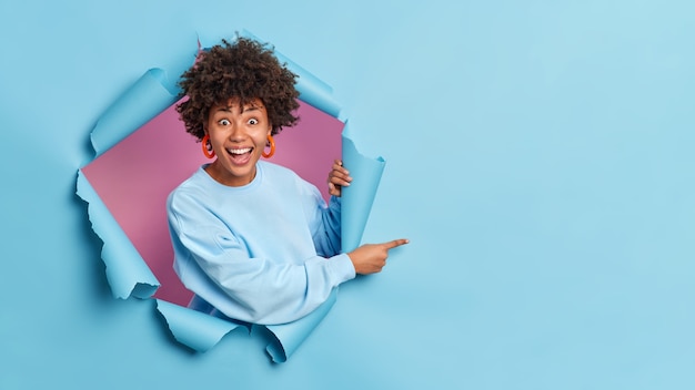 Фото Веселая удивленная женщина пробивает бумажную стену, демонстрирует пространство для копирования, а синяя стена дает советы, показывает рекламу на пустом месте, носит повседневный свитер и серьги.