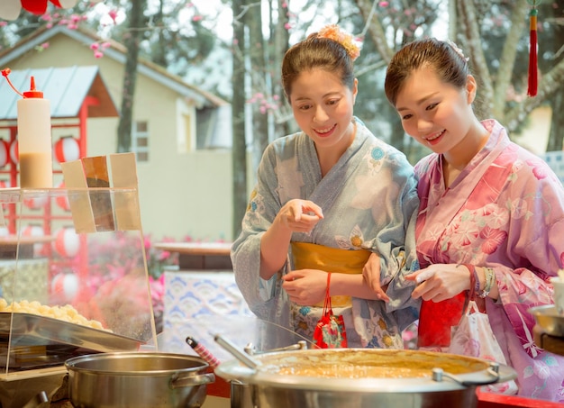 cheerful sweet women friends pointing food stall snack choosing dessert in japan and wearing traditional kimono during travel vacation.