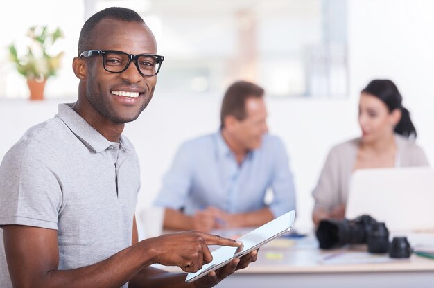 Cheerful team leader. Handsome young African man working on digital tablet and smiling while two people working on background