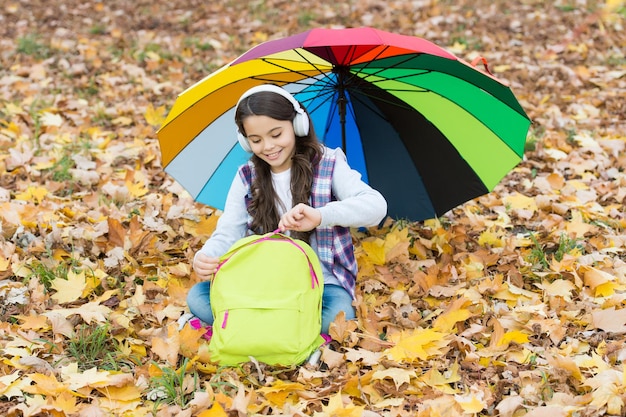 Cheerful teen child relax in fall forest or park while listening music in earphones near school backpack under colorful umbrella back to school