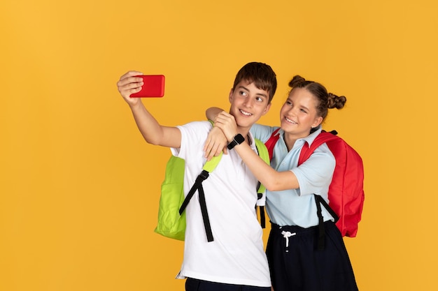 Веселые европейские мальчик и девочка с рюкзаками обнимаются и делают селфи на смартфоне