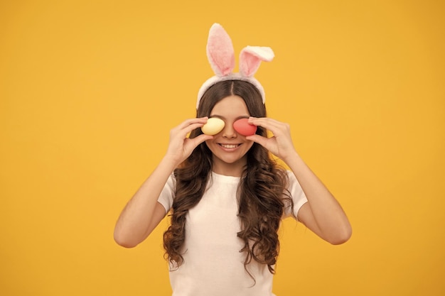 Cheerful teen girl in bunny ears hold eggs on yellow background easter