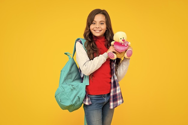 Cheerful teen girl in casual clothes holding school bag and toy bear on yellow background education