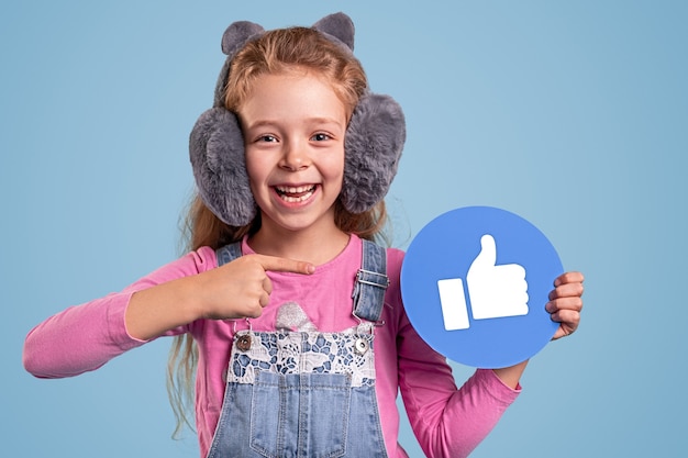 Cheerful teen girl in casual outfit and fluffy earmuffs pointing at thumb up