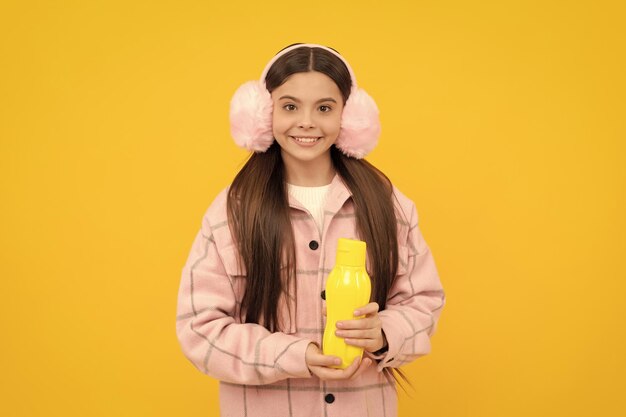 Cheerful teen girl in earmuffs with sport water bottle on yellow background drink