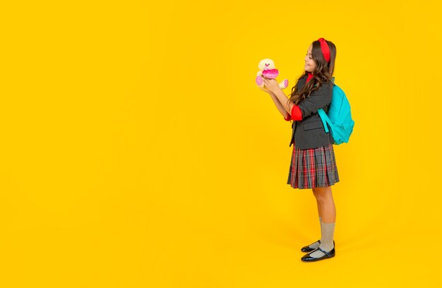 Cheerful teen girl in uniform with school bag holding toy on yellow background, september.