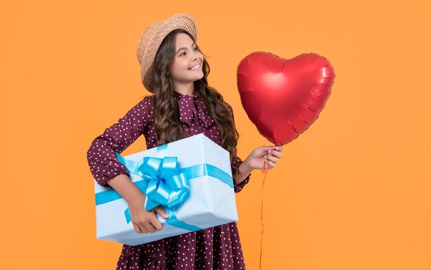 Cheerful teen kid with red heart balloon and present box on yellow background