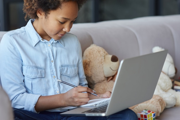 Photo cheerful teen schoolgirl using laptop making notes during online lesson while sitting on the sofa