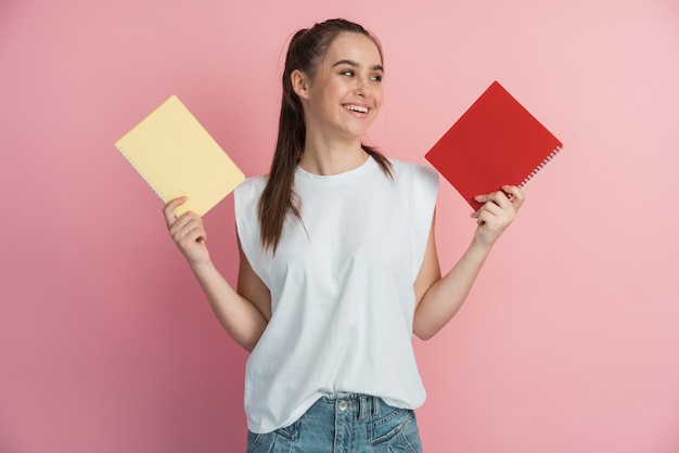 Cheerful teen woman smiling and showing laptops to camera while doing homework on pink background. d.