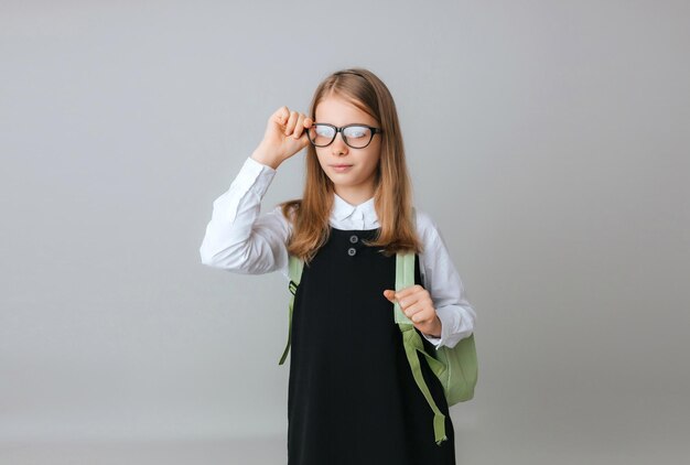 교복을 입은 111213세의 쾌활한 십대 소녀는 회색 배경 학교 패션에 배낭을 착용합니다
