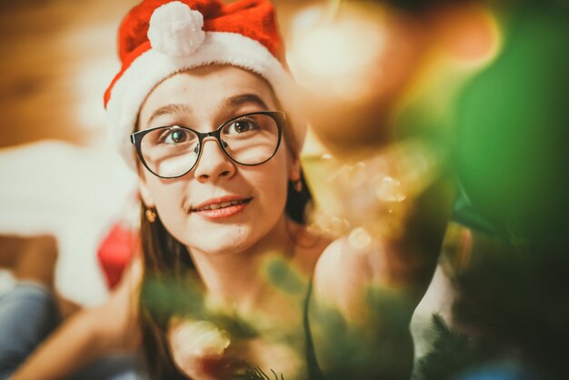 Photo cheerful teenage girl wearing glasses and santa hat decorates christmas tree