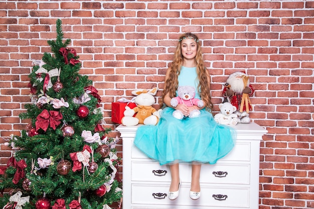 Cheerful teenage long haired blonde sit on a white nightstand near christmas tree, toothy smile and looking at camera. Studio shot