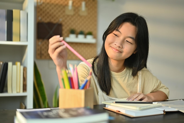 cheerful teenaged girl doing homework writing in notebook while sitting at table in living room