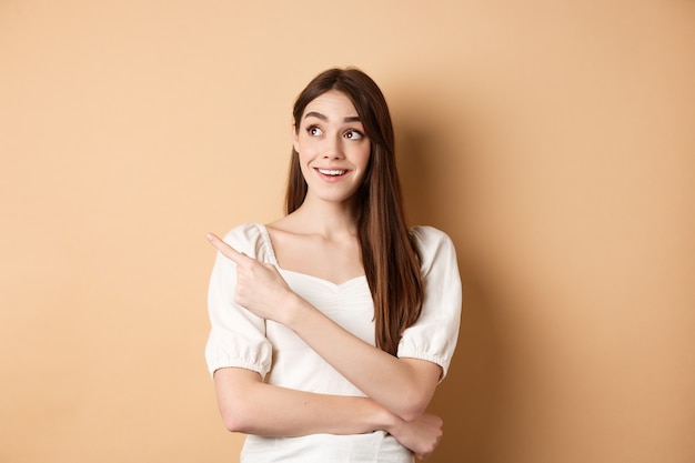 Cheerful tender girl in white dress checking out promo offer, looking and pointing at upper left corner with dreamy smile, standing on beige background