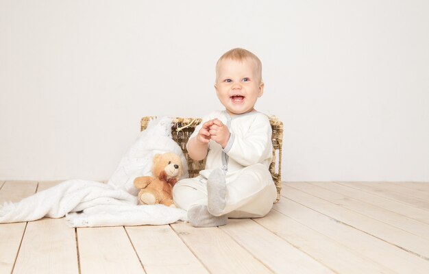 cheerful toddler in white cloth sitting with blanket and bear smiling at camera on white background.