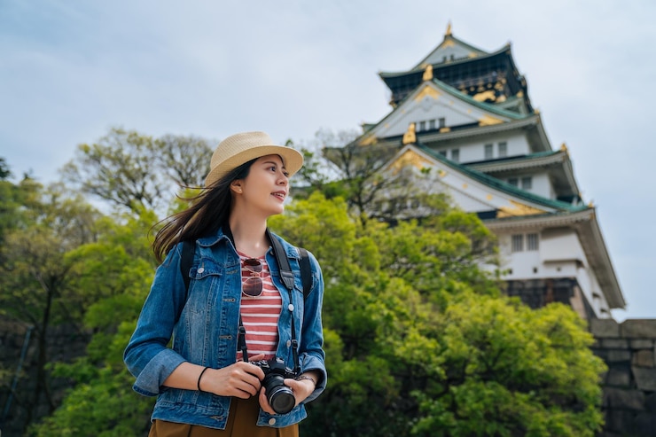 Photo 陽気な観光写真家の趣味は、日本の都市を観光する一眼レフカメラで旅行するのが大好きです。大阪宮殿の前に立つ若い魅力的な旅行者。女性の楽しい独立旅行。