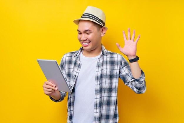 Cheerful traveler tourist asian man in casual clothes hat using a digital tablet waving his hand gets a video call on yellow background passenger traveling on weekends air flight journey concept