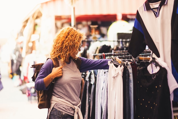 Viaggiatore allegro giovane donna riccia bionda che guarda e sceglie i vestiti al mercato usato durante le vacanze alternative
