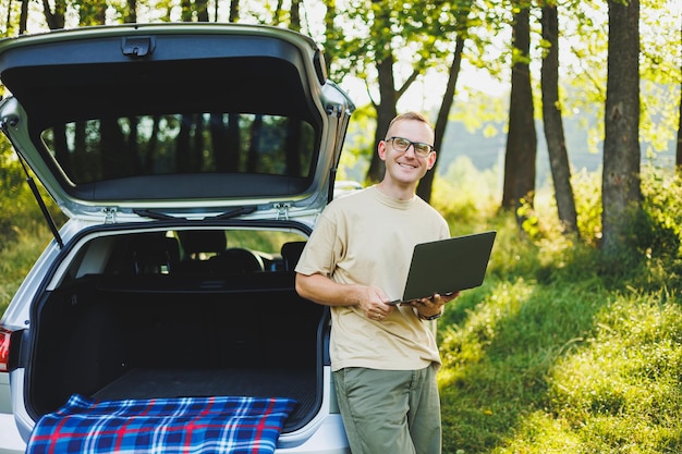 Cheerful traveler young man sitting in open trunk of car working with laptop outdoors concept of business travel vacation