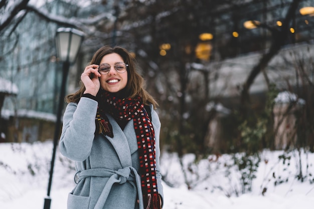 Cheerful trendy adult beautiful female in coat jacket and eyeglasses at street with snow