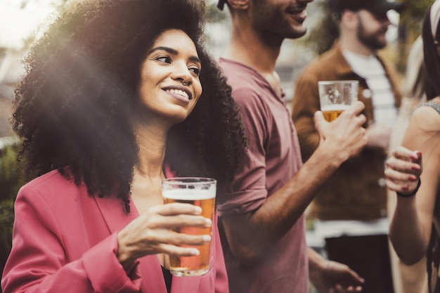 Cheerful trendy brazilian girl in a students rooftop party\
holding a glass of beer
