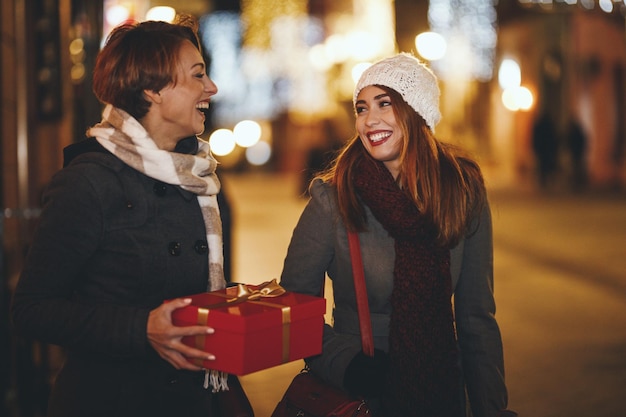 陽気な2人の若い女性がクリスマスの夜に街の通りで楽しんでいます。彼らは笑って、これからの休日のためにプレゼントを買っています。