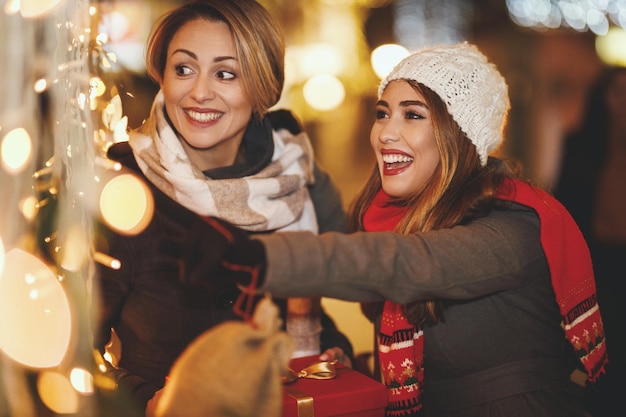 陽気な2人の若い女性がクリスマスの夜に街の通りで楽しんでいます。彼らは笑って、これからの休日のためにプレゼントを買っています。