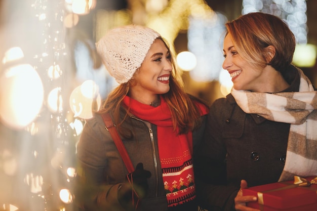 陽気な2人の若い女性がクリスマスの夜に街の通りで楽しんでいます。彼らは笑って、これからの休日のためにプレゼントを買っています。