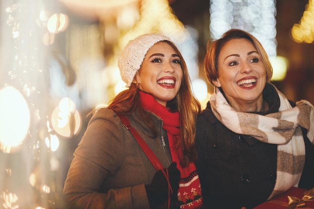 陽気な2人の若い女性がクリスマスの夜に街の通りで楽しんでいます。彼らは笑って、これからの休日のためにプレゼントを買っています。