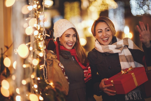 陽気な2人の若い女性がクリスマスの夜に街の通りで楽しんでいます。彼らは笑って、これからの休日のためにプレゼントを買っています。