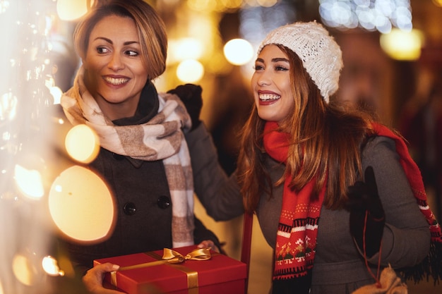 陽気な2人の若い女性がクリスマスの夜に街の通りで楽しんでいます。彼らは笑って、最愛の人へのプレゼントを買っています。