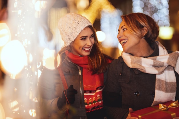 陽気な2人の若い女性がクリスマスの夜に街の通りで楽しんでいます。彼らは笑って、最愛の人へのプレゼントを買っています。