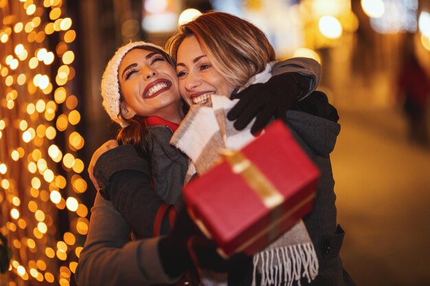 Cheerful two young women are having fun in the city street at the Christmas night. They are laughing, hugging and giving presents to each other.