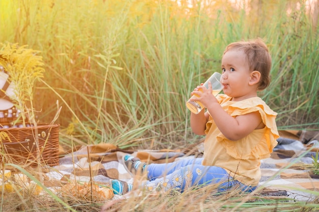 Un'allegra ragazza di due anni con una maglietta gialla e pantaloni blu sta bevendo succo in natura un bambino sotto i raggi del sole al tramonto in natura