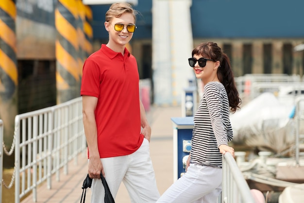 Cheerful wealthy young couple in sunglasses standing on pier in port and waiting for vessel