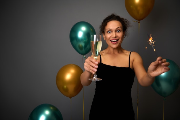 Cheerful woman in black evening dress holds a glass of champagne and sparklers, poses against gray background with beautiful decoration from gold and green air balloons. Christmas, Birthday concept