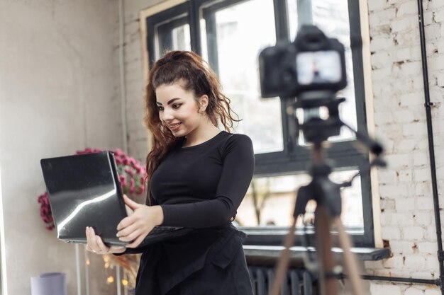 cheerful woman blogger recording video blog on camera with laptop in her hands indoors