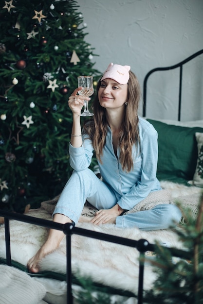 Cheerful woman in blue sleepwear drinks alcohol on the bed