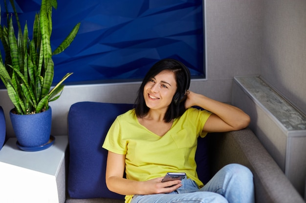 Cheerful woman in casual clothes with headphones and smartphone, listening to podcast on sofa at home.