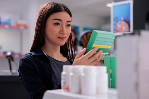 쾌활한 여성 고객이 약국에서 약을 쇼핑하는 동안 의약품 전단지를 보고 있는 약 패키지를 들고 있고, 고객은 건강한 면역 체계를 위한 보충제와 비타민을 구입하고 있습니다. 의약품 지원