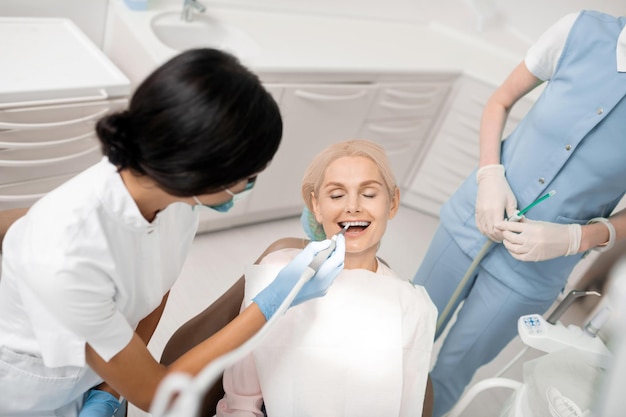 Cheerful woman closing her eyes and opening her mouth getting her teeth treatment in dental clinic