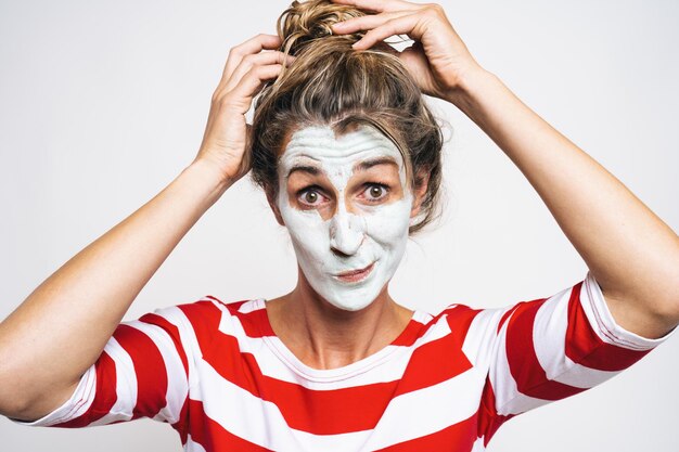 Cheerful woman correcting hair bun, has clay mask on face,\
enjoys softness, has beauty treatments at home, on gray background.\
skin care concept image