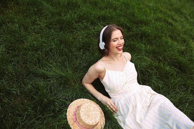 쾌활한 여자 드레스와 헤드폰은 음악을 듣고 야외에서 멀리 보는 동안 잔디에 누워