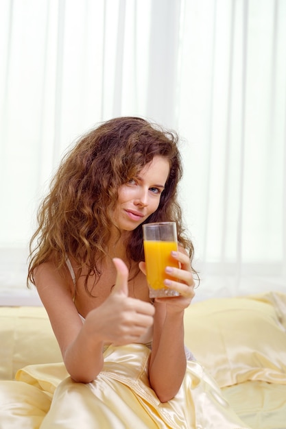 Веселая женщина пьет апельсиновый сок, сидя на кровати у себя дома